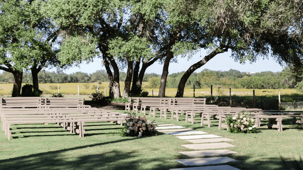 Wooden benches are arranged outdoors on grass for a ceremony at one of the charming wedding venues in Austin, with floral arrangements and stone pavers leading to a focal point, surrounded by trees and open fields.