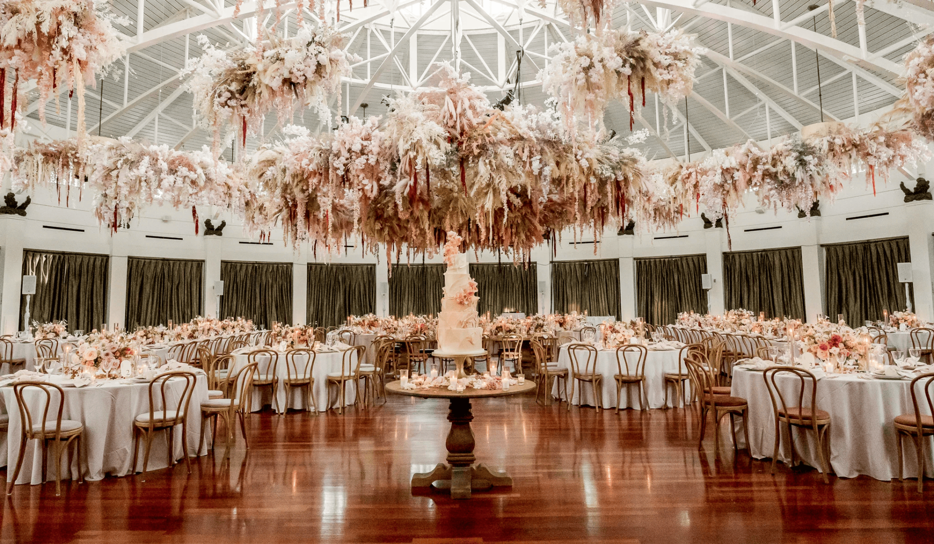 Elegant banquet hall among top New Orleans wedding venues, with round tables set for an event, a tall cake on a central table, and abundant floral arrangements hanging from the ceiling.
