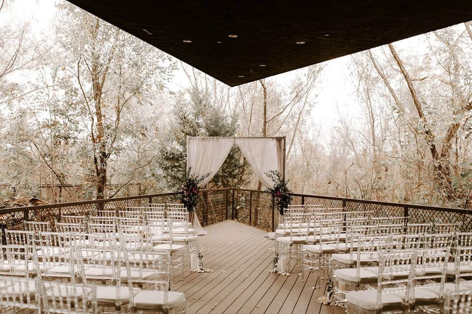 Rows of white chairs face a simple wedding arch with white drapery and floral arrangements on a wooden deck surrounded by trees, offering the perfect setting for those searching for wedding venues in Fort Worth.