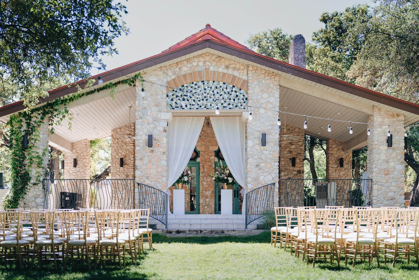 Stone pavilion with white drapes, floral arrangements, and rows of wooden chairs arranged for an outdoor ceremony—an elegant choice among San Antonio wedding venues.