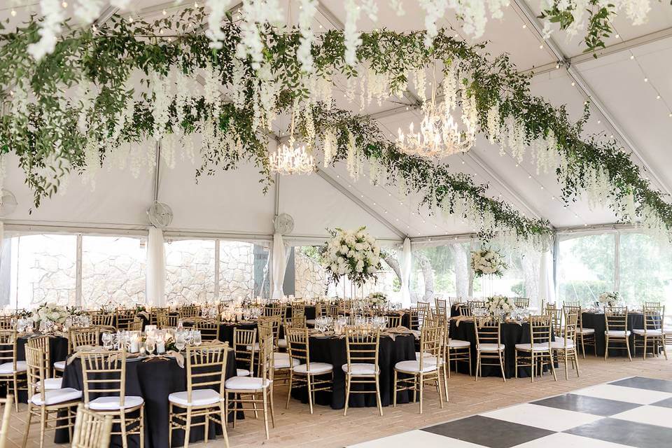 Elegant event tent with chandeliers, hanging greenery, round tables covered in black tablecloths, gold chairs, and a black-and-white checkered dance floor—perfect inspiration for wedding venues in Austin.