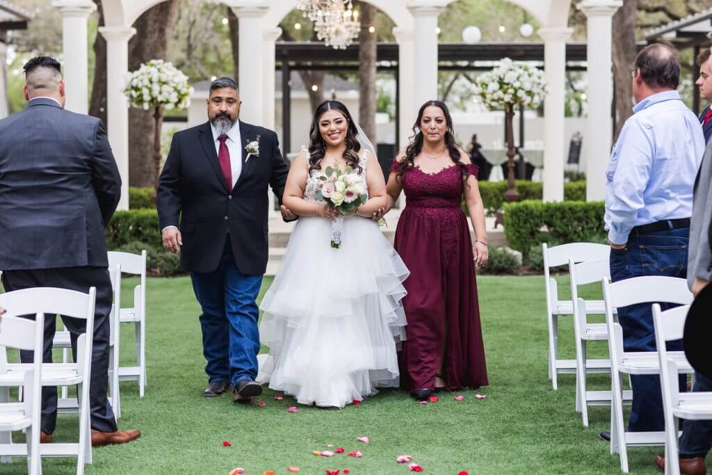 Bride walking down the aisle arm in arm with her parents at an outdoor South Texas wedding ceremony framed by white columns and floral arrangements