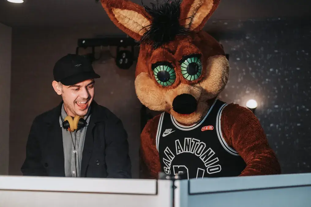 Toast DJ laughing behind the booth at a San Antonio event as the Spurs Coyote leans in beside him